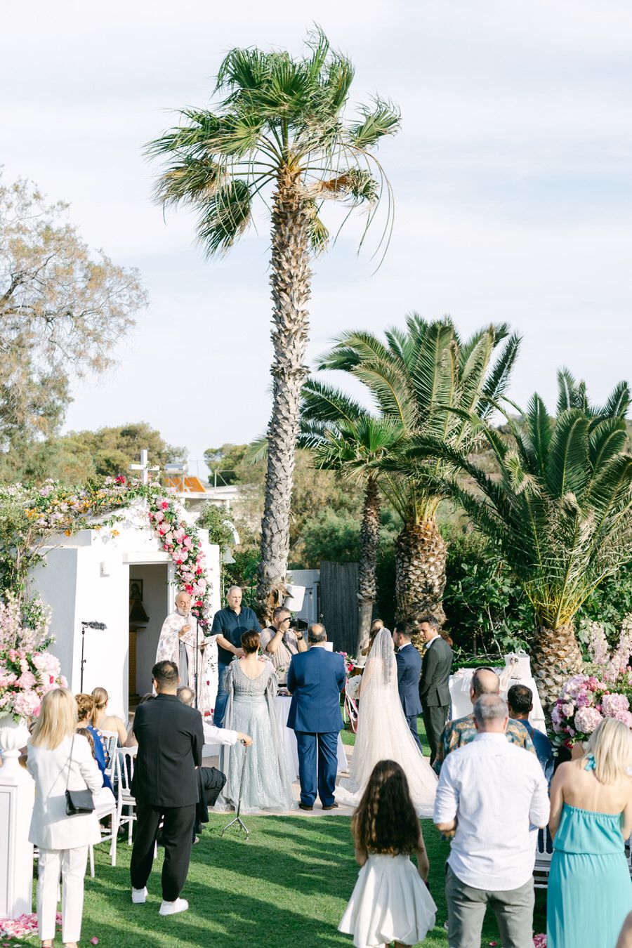 Palm tree-lined outdoor wedding ceremony with guests seated on white chairs.