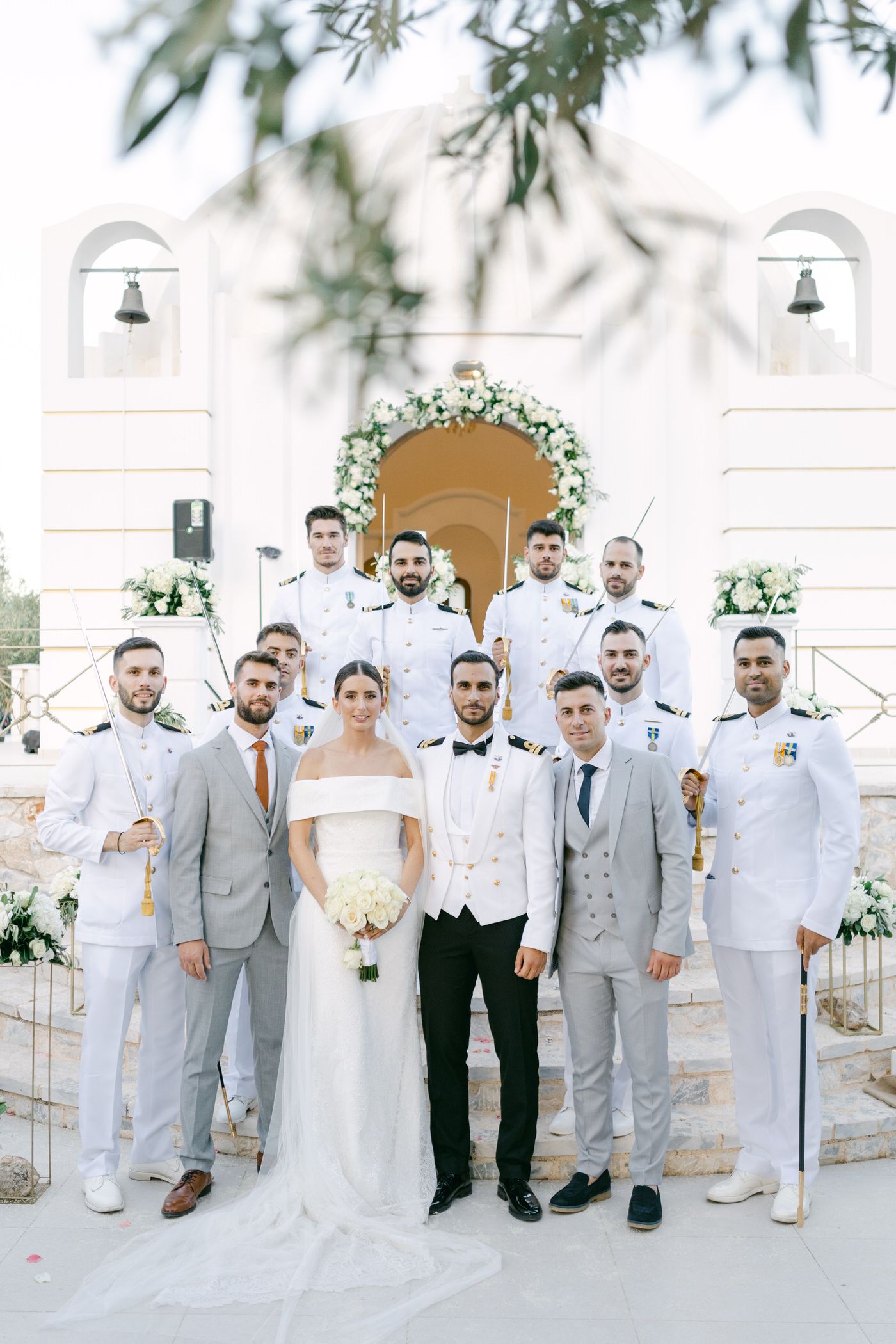 Wedding party dressed in shades of white and gray poses together in front of a church entrance with decorative floral arch.