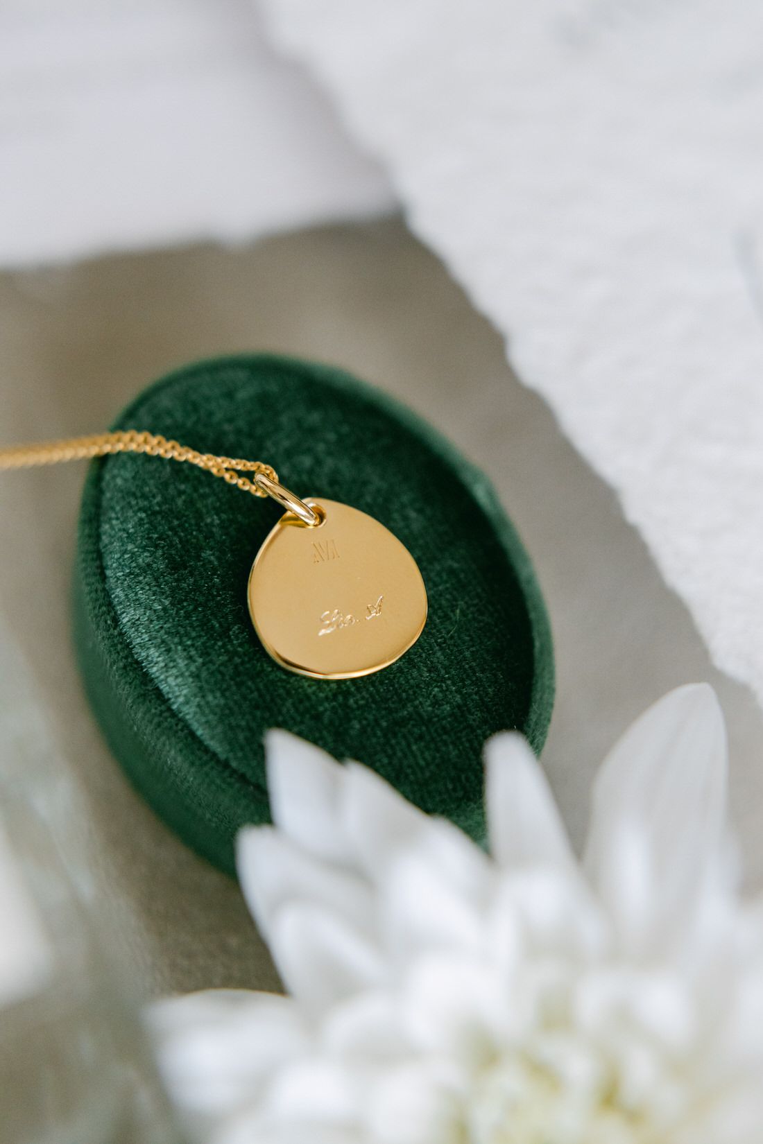 A delicate gold disc necklace rests on an emerald velvet cushion beside a white flower petal.
