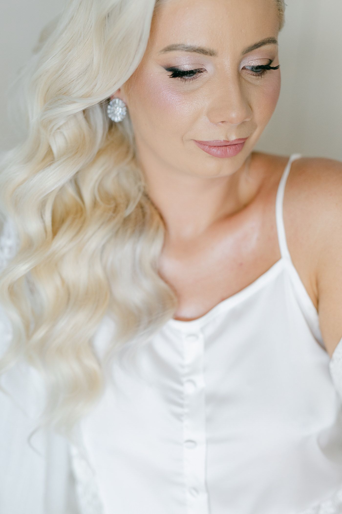 Elegant bridal portrait with platinum blonde waves, white satin top and delicate makeup in soft natural lighting.