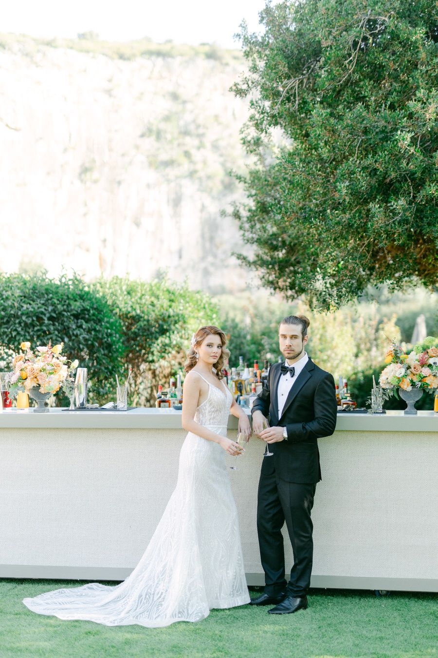 Elegant wedding couple posing by white bar with floral arrangements and mountain backdrop.