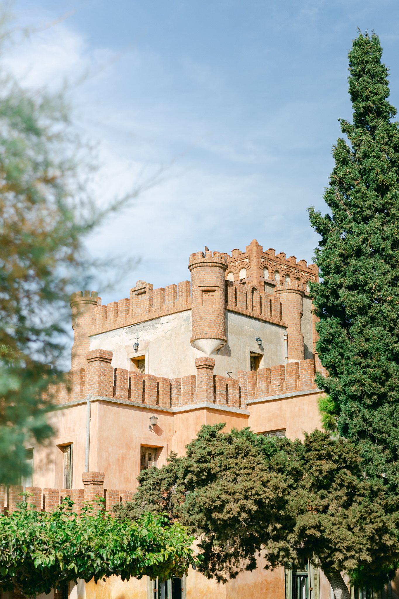 Ancient castle-like building with crenellated towers and terracotta walls surrounded by green cypress trees under blue sky.
