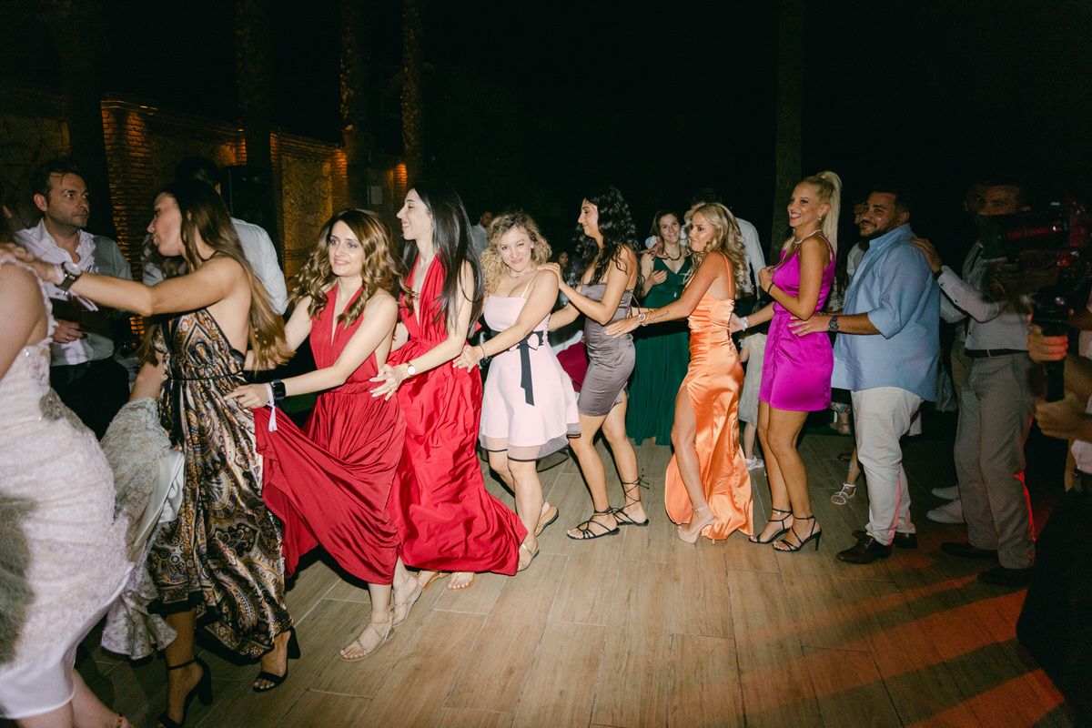 A line of dancers in formal attire performing a conga line dance on a wooden dance floor at a celebration event.