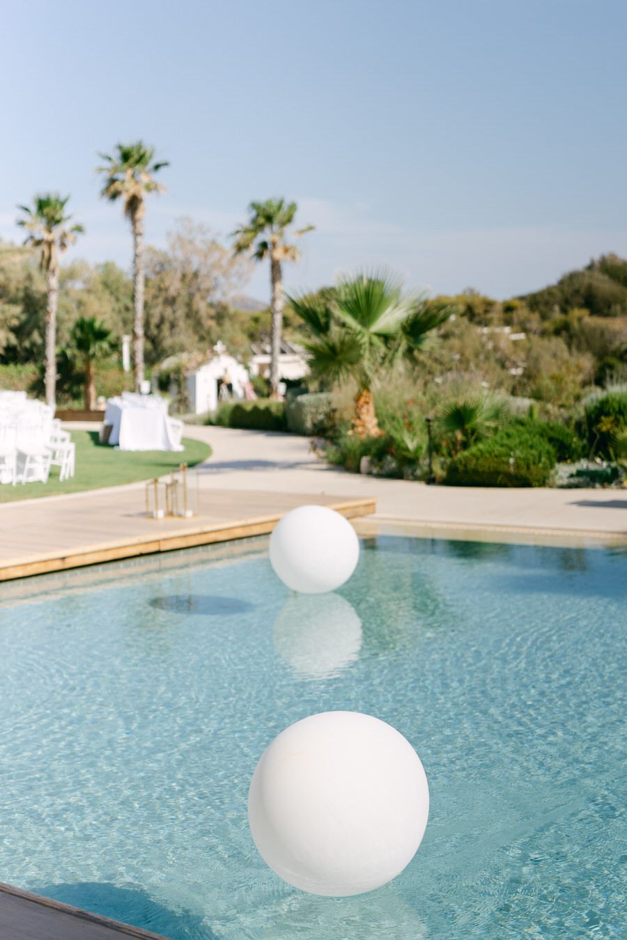 White decorative spheres floating in pool with palm trees and wedding setup in background.