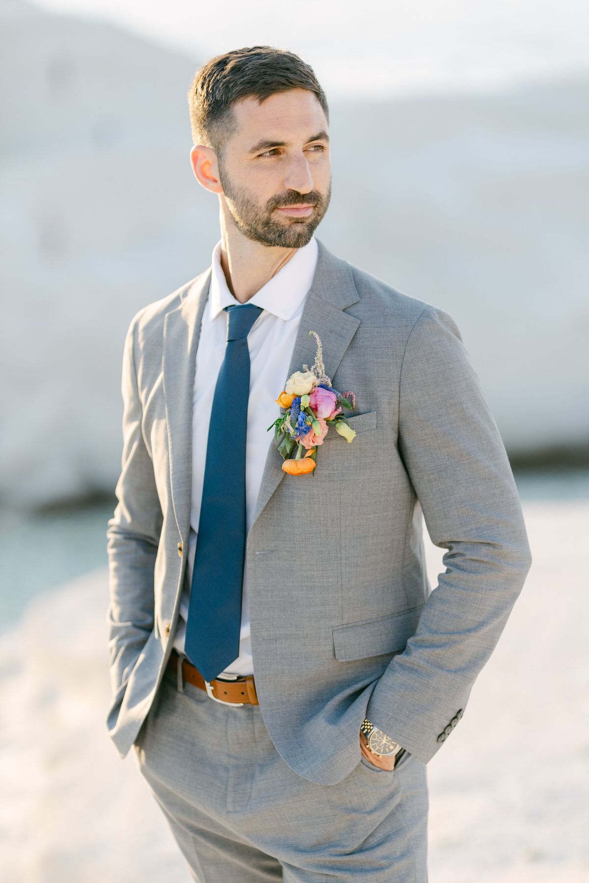 A groom in a light gray suit with navy tie and colorful floral boutonniere poses outdoors with mountains in background.
