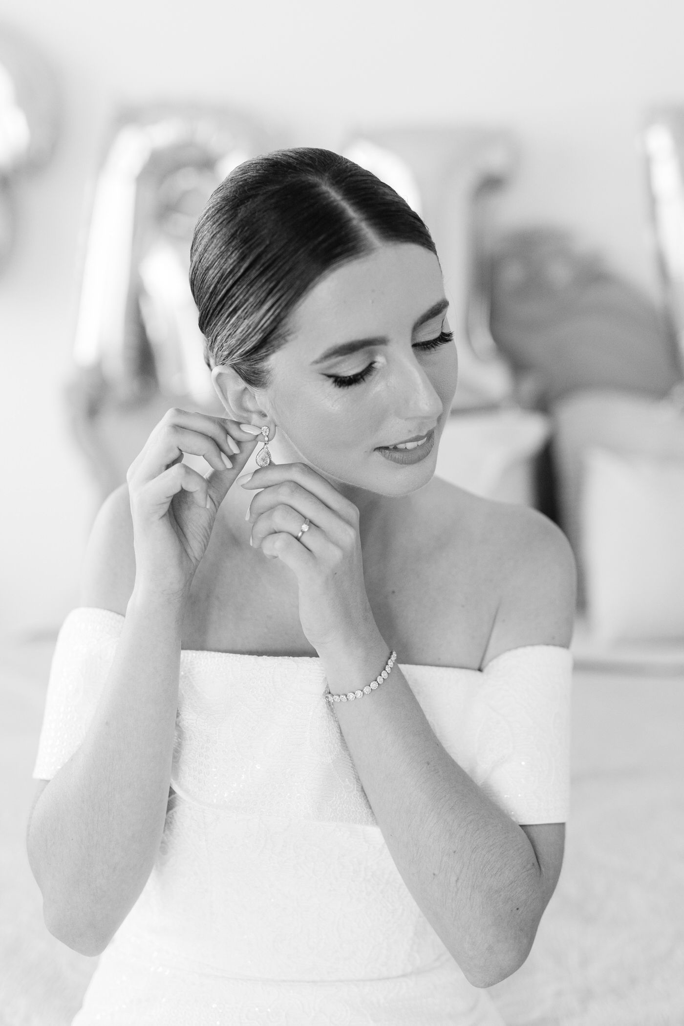Black and white photo of someone putting on earrings while wearing an elegant off-shoulder white dress and bracelet.