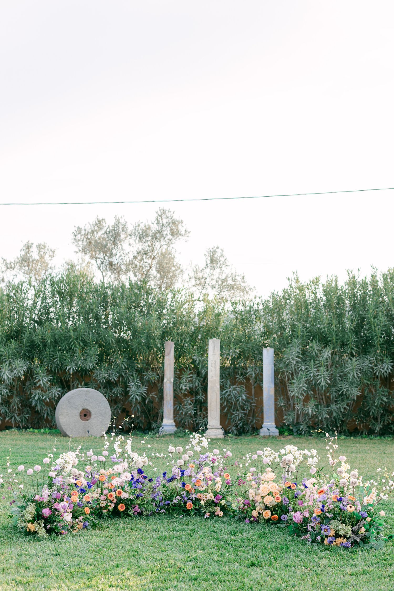 Elegant outdoor wedding ceremony setup with white pillars and floral arrangements against a green garden backdrop.