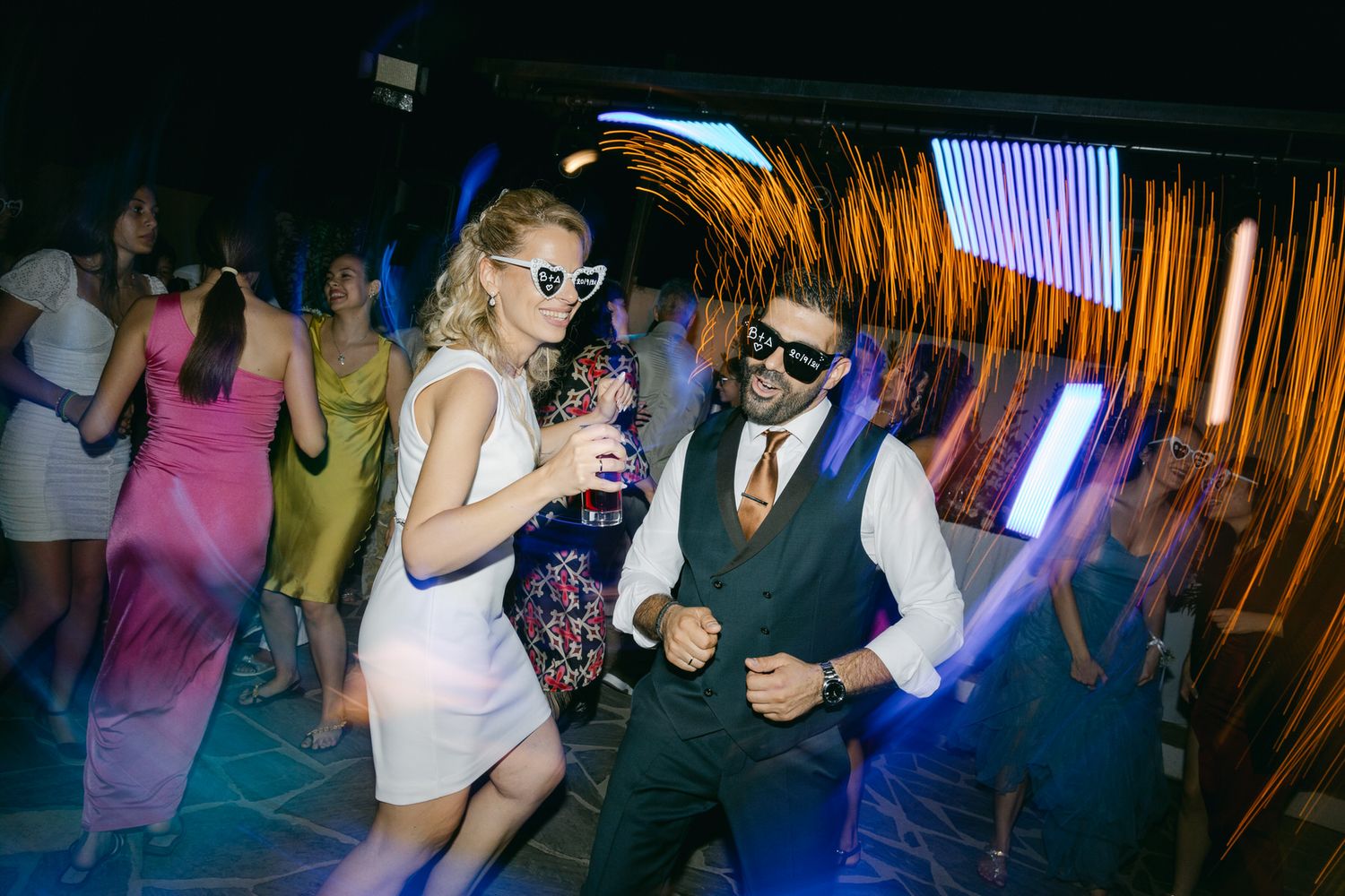 Wedding guests dance energetically in sunglasses on a colorful dance floor at an evening reception.