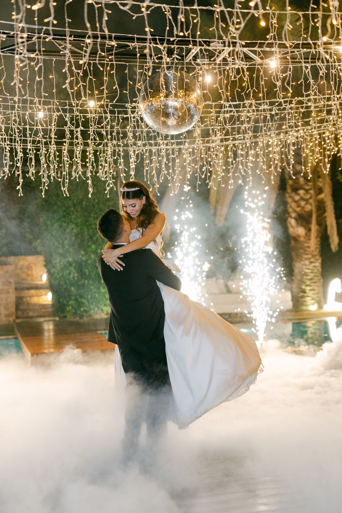 A dreamy wedding couple dances amid fog effects under twinkling string lights and disco balls in an enchanted outdoor setting.