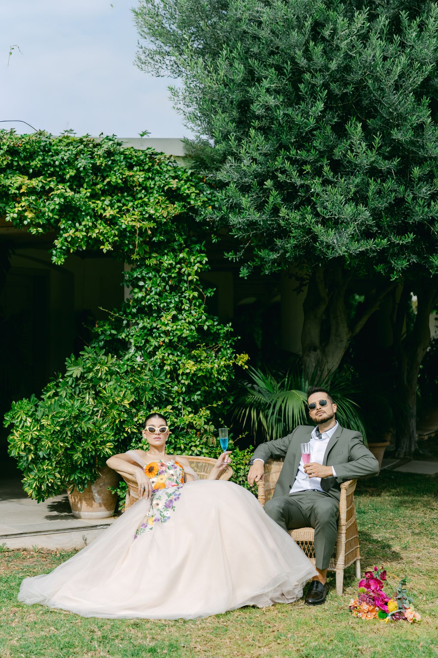 Couple in sunglasses relaxing in garden setting with lush greenery and a flowing white dress. 2026 2027 γάμος