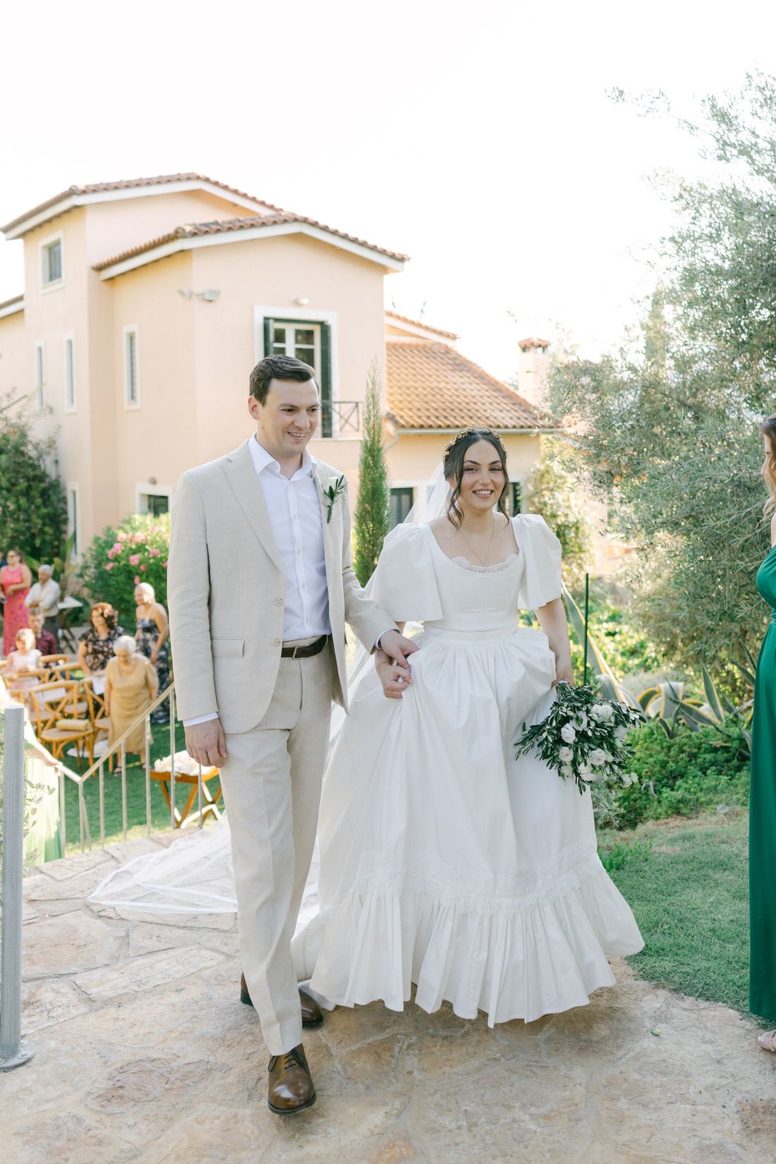 A couple walks down a garden path in wedding attire with a peach-colored Mediterranean style house in the background.
