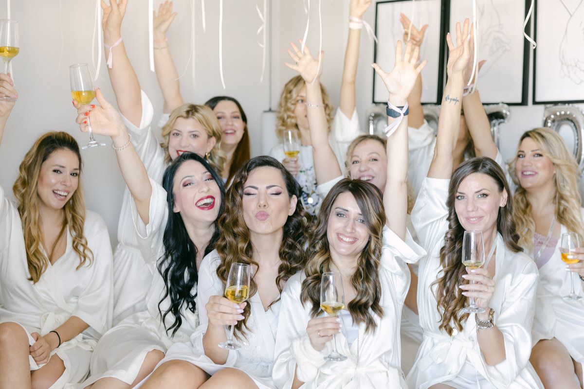 Group wearing white robes raise champagne glasses in celebration during a bridal party gathering.