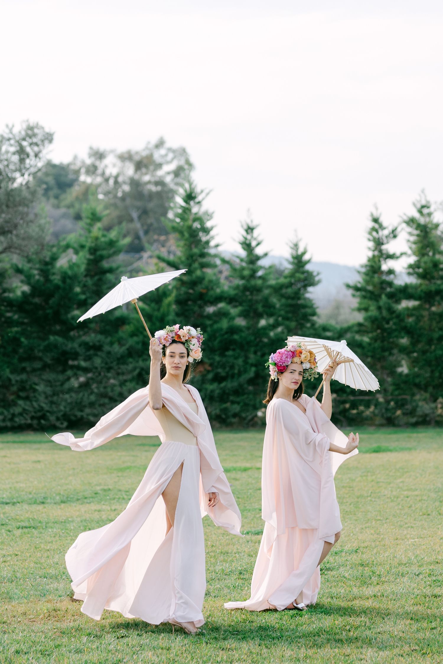 Two women in flowing white dresses and floral crowns dance with parasols in a grassy field near evergreen trees.