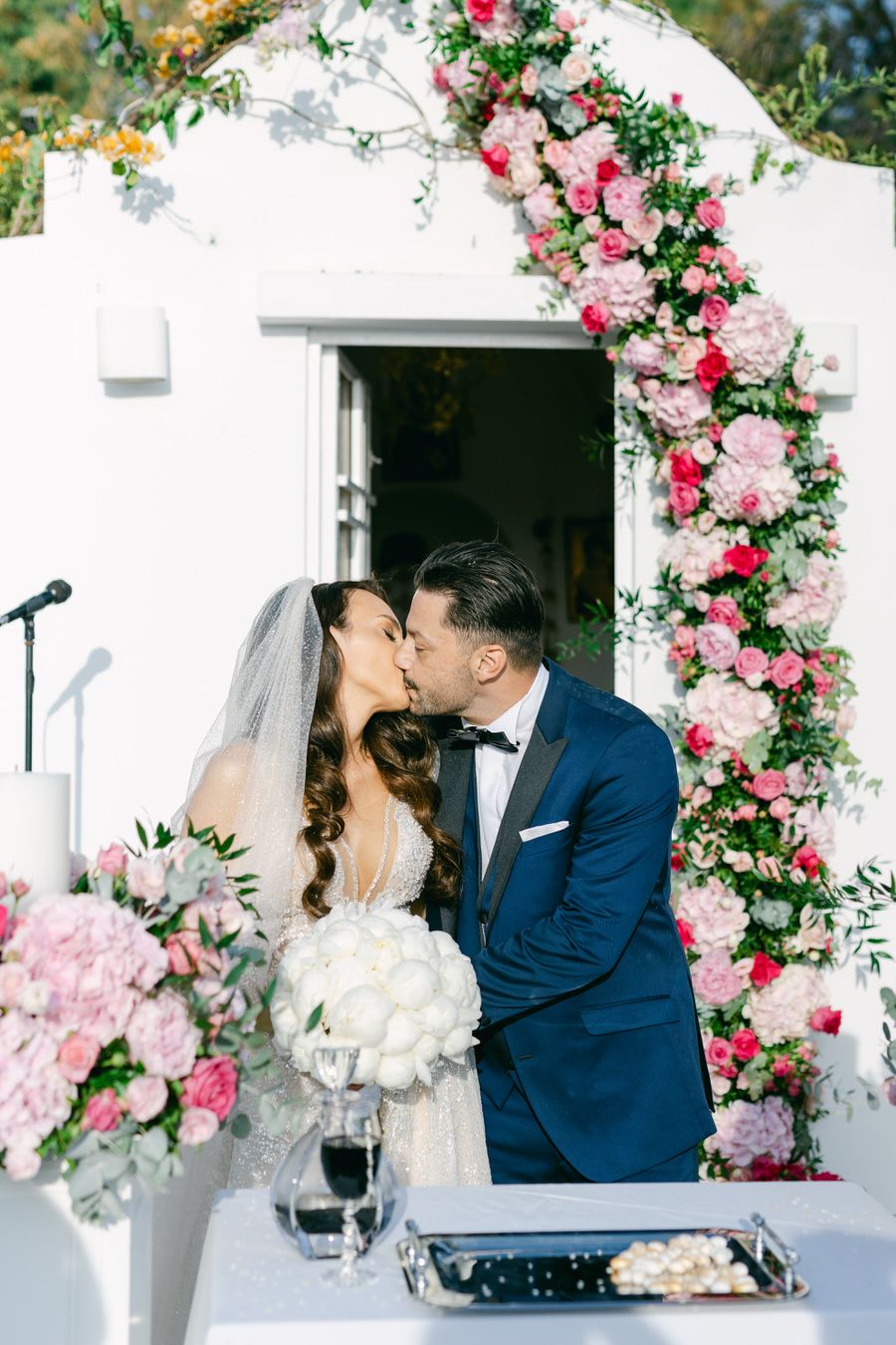 Romantic wedding kiss under an arch decorated with pink roses and greenery.