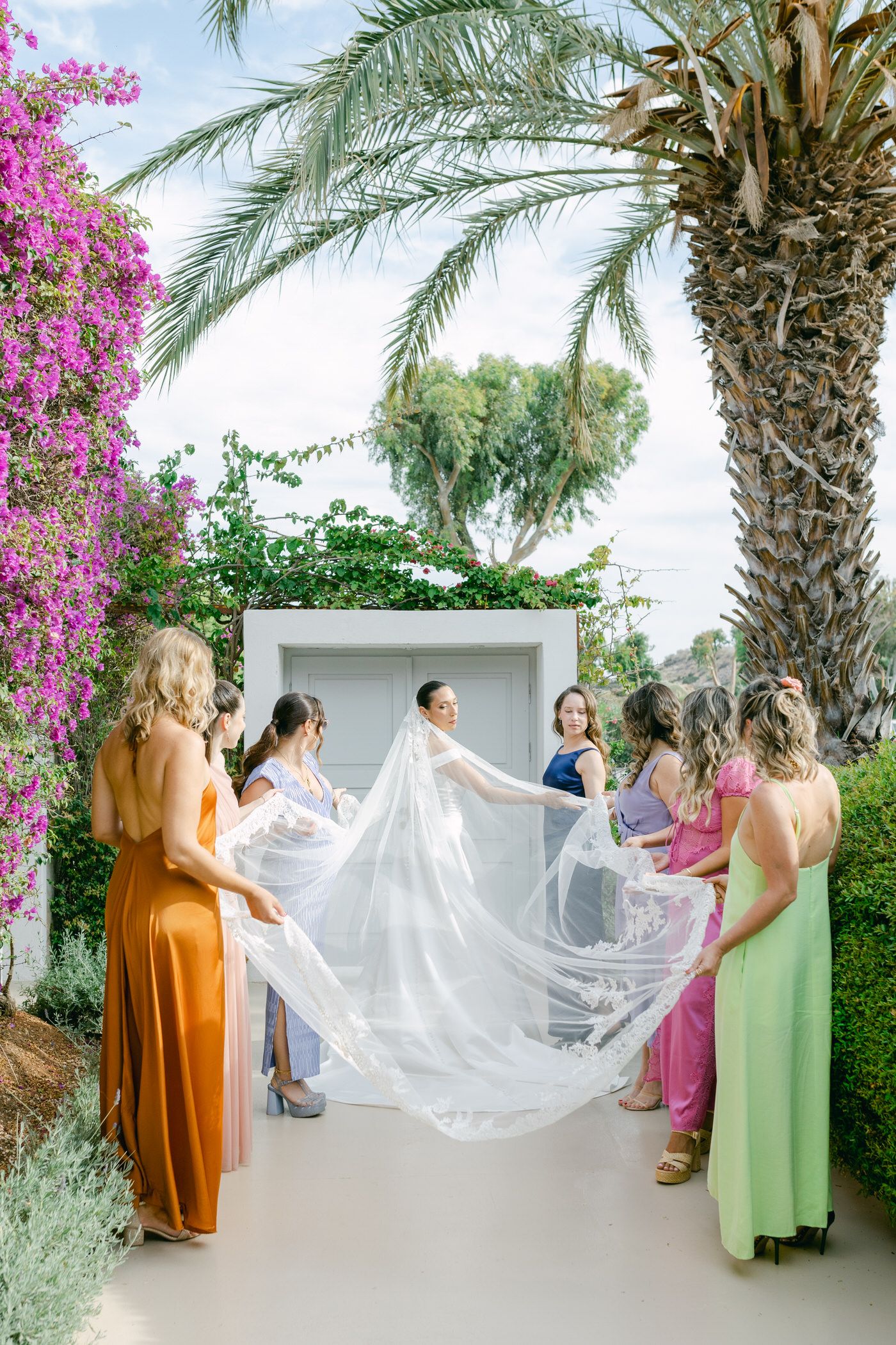 Bridesmaids in colorful dresses hold a flowing white veil under swaying palm trees and vibrant bougainvillea flowers at a resort.