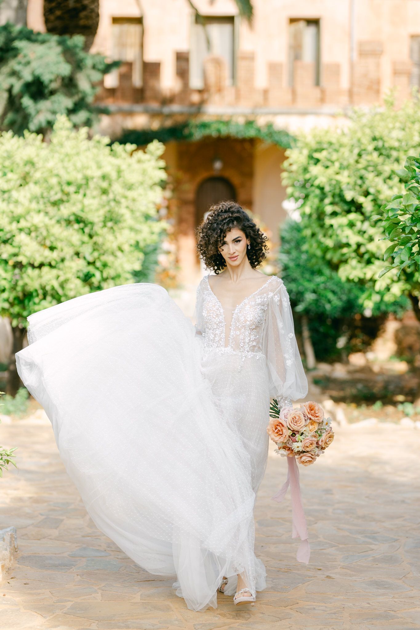 A bride in a flowing white lace gown with long sleeves holds a peach bouquet while walking in a garden courtyard.