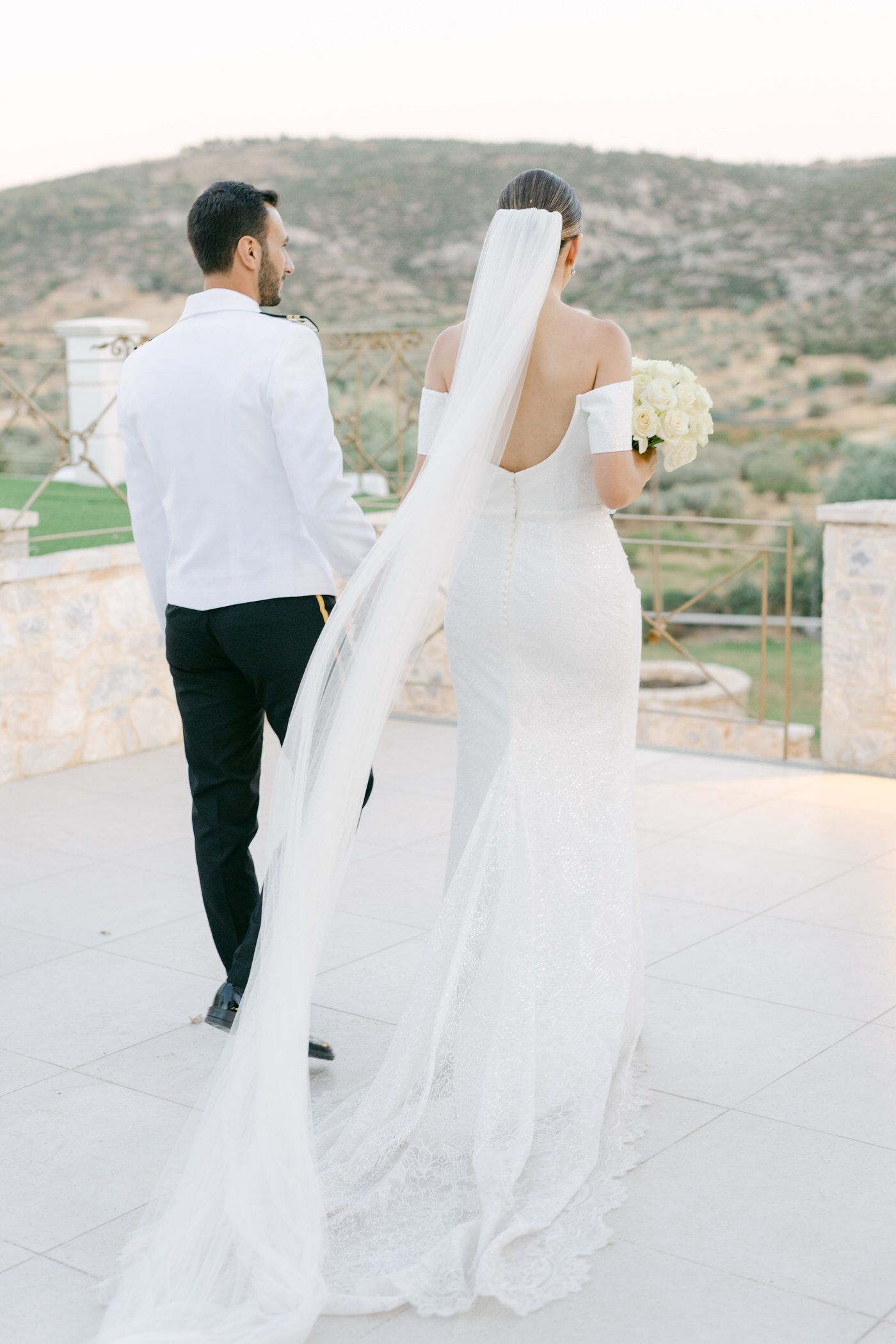 A bride in a white off-shoulder gown with flowing veil stands with groom on a terrace overlooking mountainous landscape.
