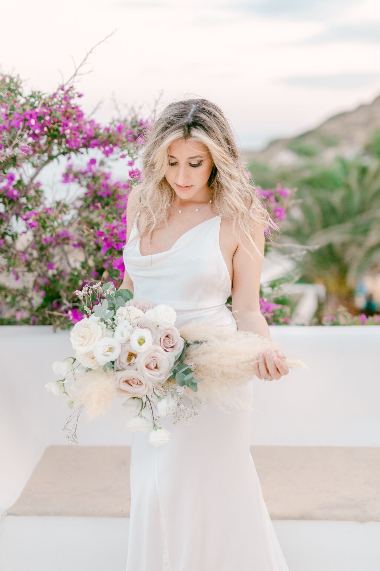 A bride in a white dress holds a romantic bouquet of blush roses against a backdrop of vibrant bougainvillea flowers.
