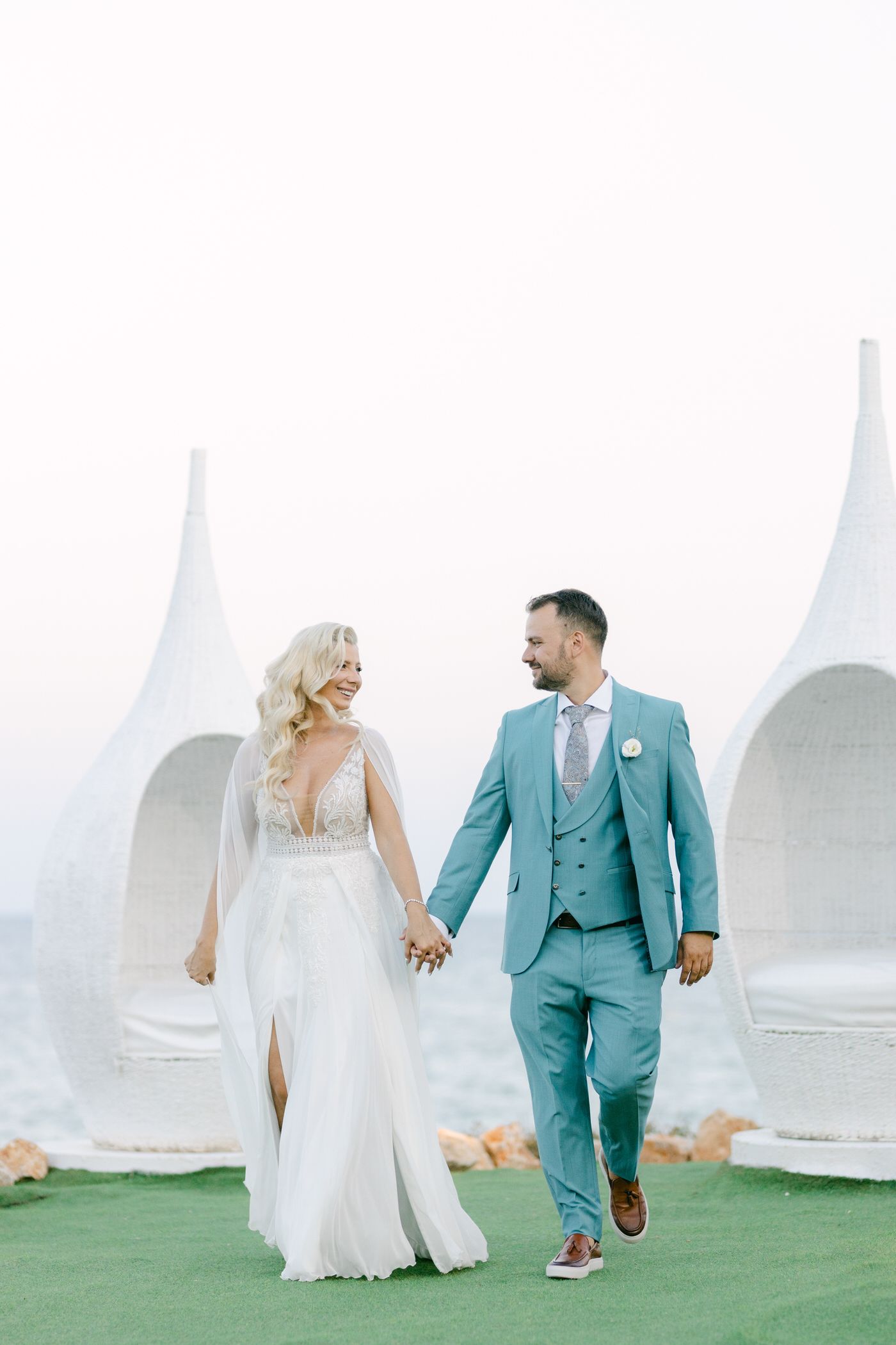 A couple walks hand in hand on green grass, with the bride in white dress and groom wearing a turquoise suit.