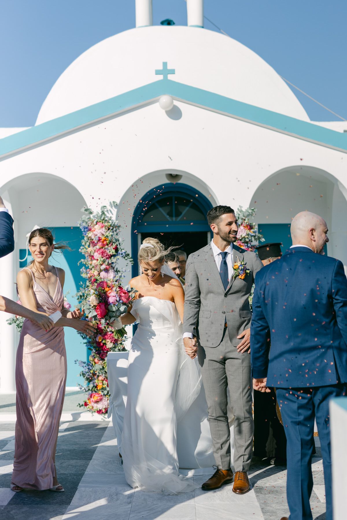 A wedding ceremony takes place outside a white Mediterranean-style church with blue accents and floral arch decorations.