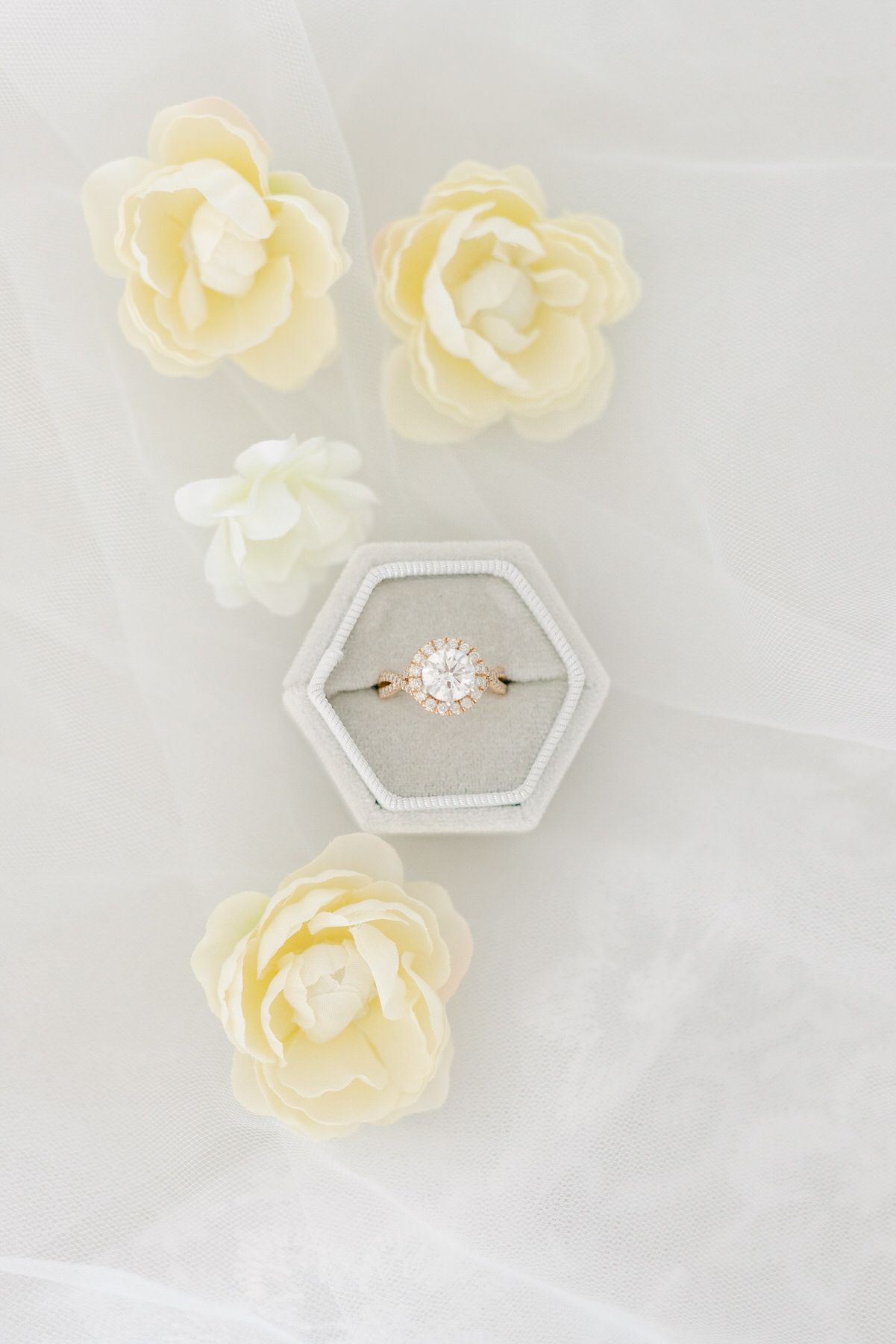 A delicate engagement ring in a hexagonal box surrounded by soft cream-colored rose petals on white tulle fabric.