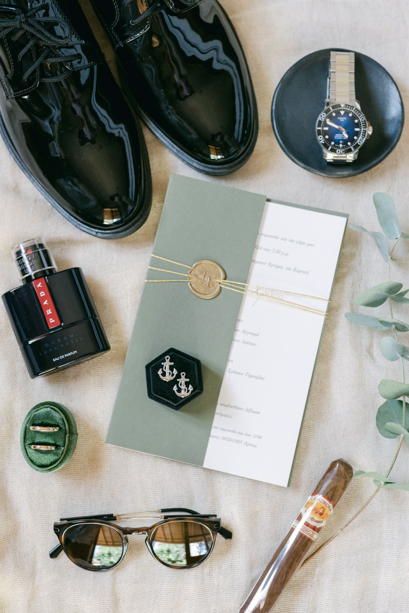 Wedding day accessories including shiny black dress shoes, watch, invitation, sunglasses, cologne, and cigar arranged on beige.