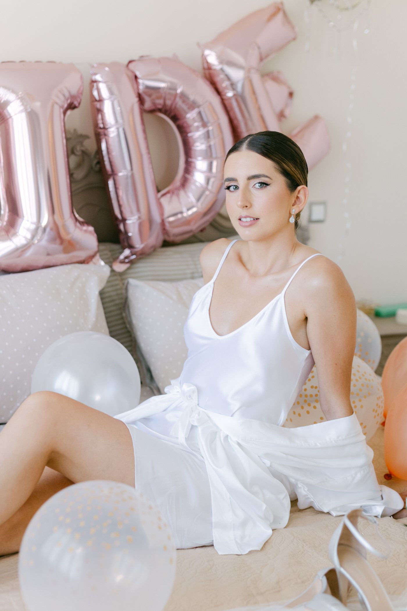 Woman in white dress poses elegantly with rose gold letter balloons and white decorative balloons in background.