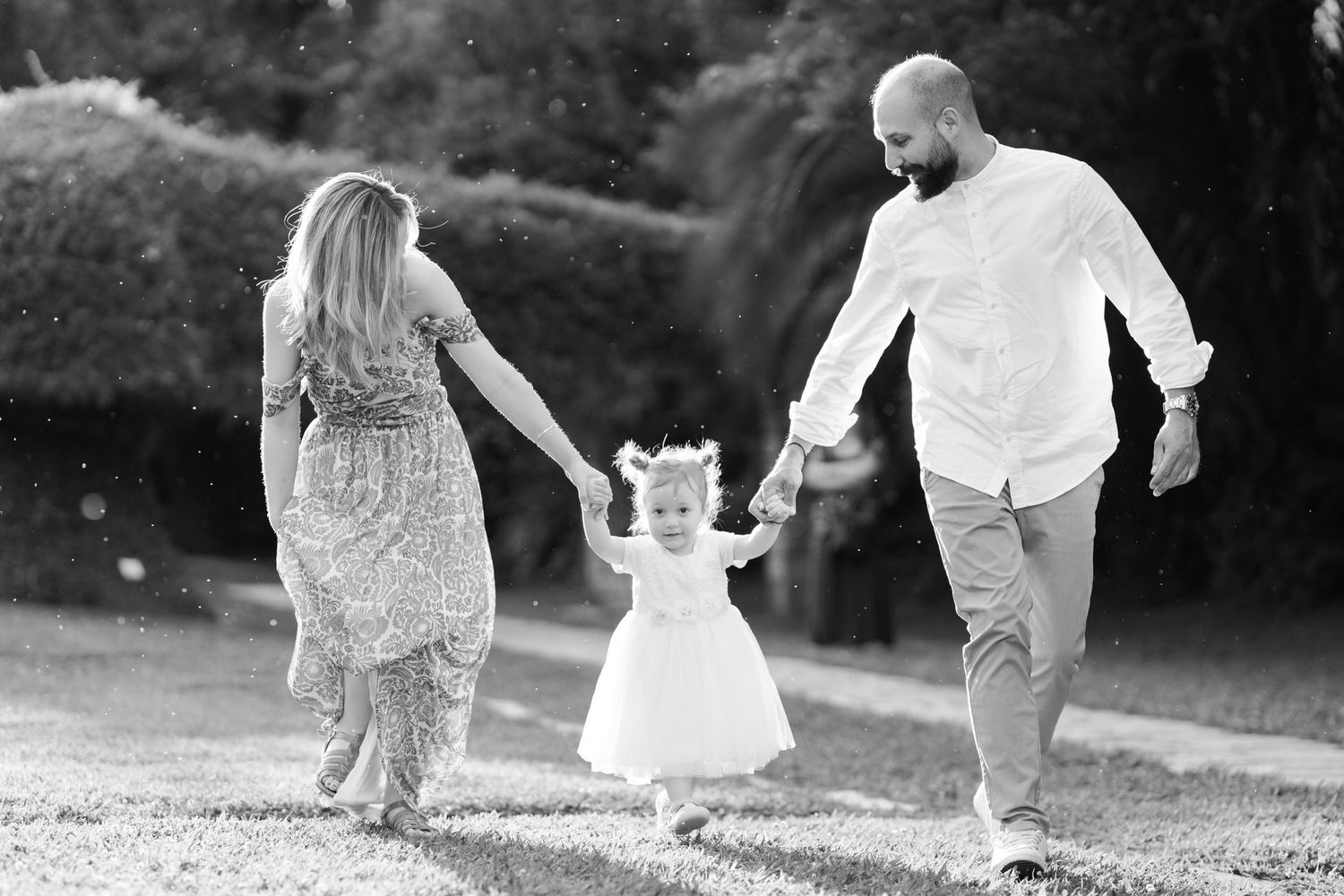 A black and white photograph shows a couple walking hand in hand with a small child in a white dress outdoors. Βάπτιση Tonia and Theodore