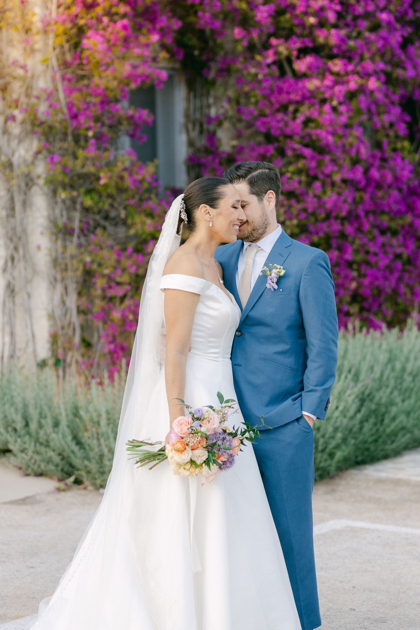 A romantic wedding moment set against a vibrant purple bougainvillea wall, with the couple sharing an intimate embrace.