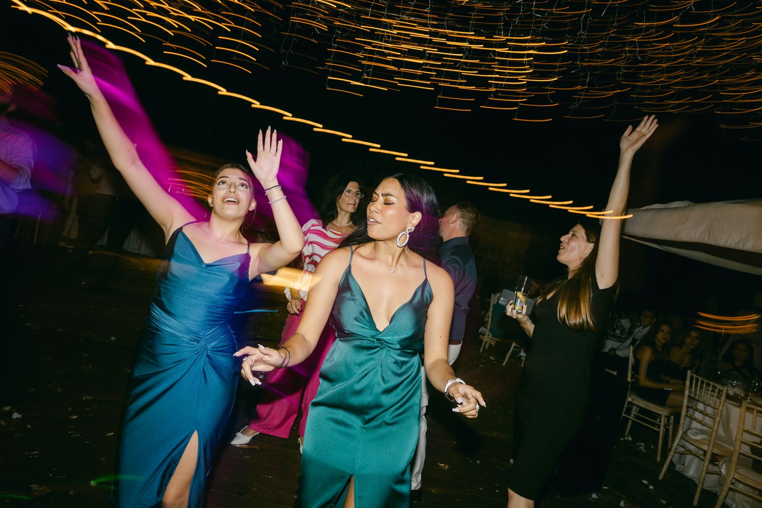 Party guests in blue dresses dance enthusiastically on a dimly lit dance floor at an evening celebration.