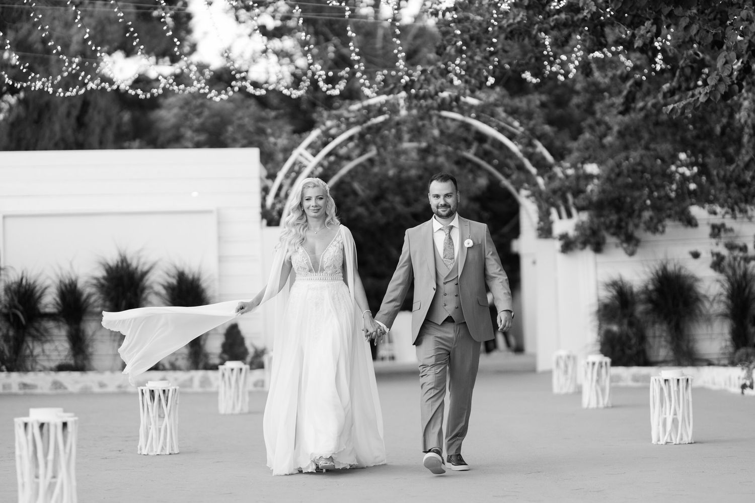 A couple walks together under a decorative garden archway at an elegant wedding venue in black and white.