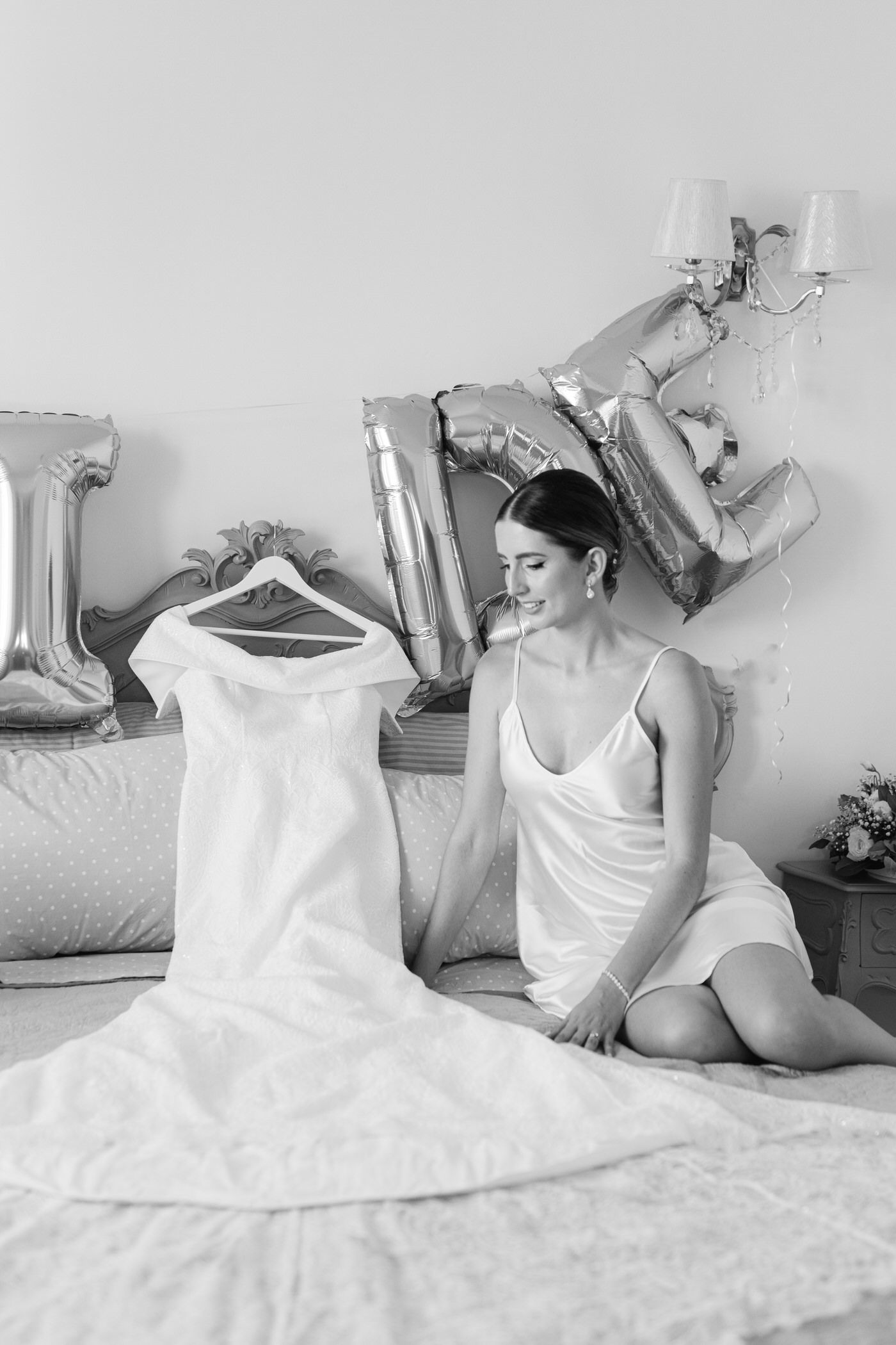 A black and white photo showing a bridal gown laid out on a bed next to someone in a white slip dress.