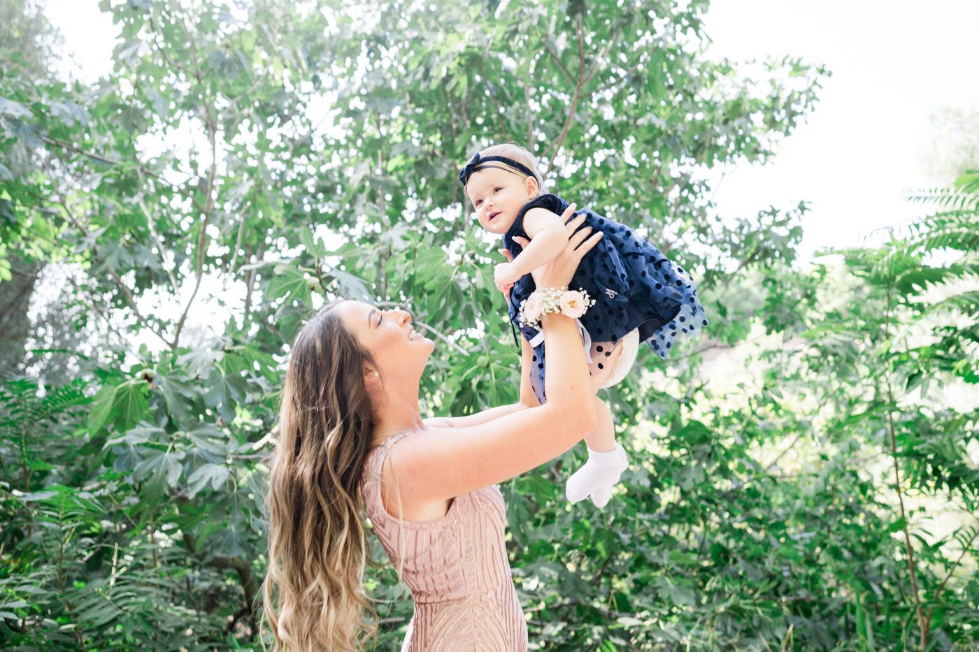 A caregiver lifts a baby wearing a navy blue outfit high into the air among green trees on a bright summer day. Βάπτιση Tonia and Theodore