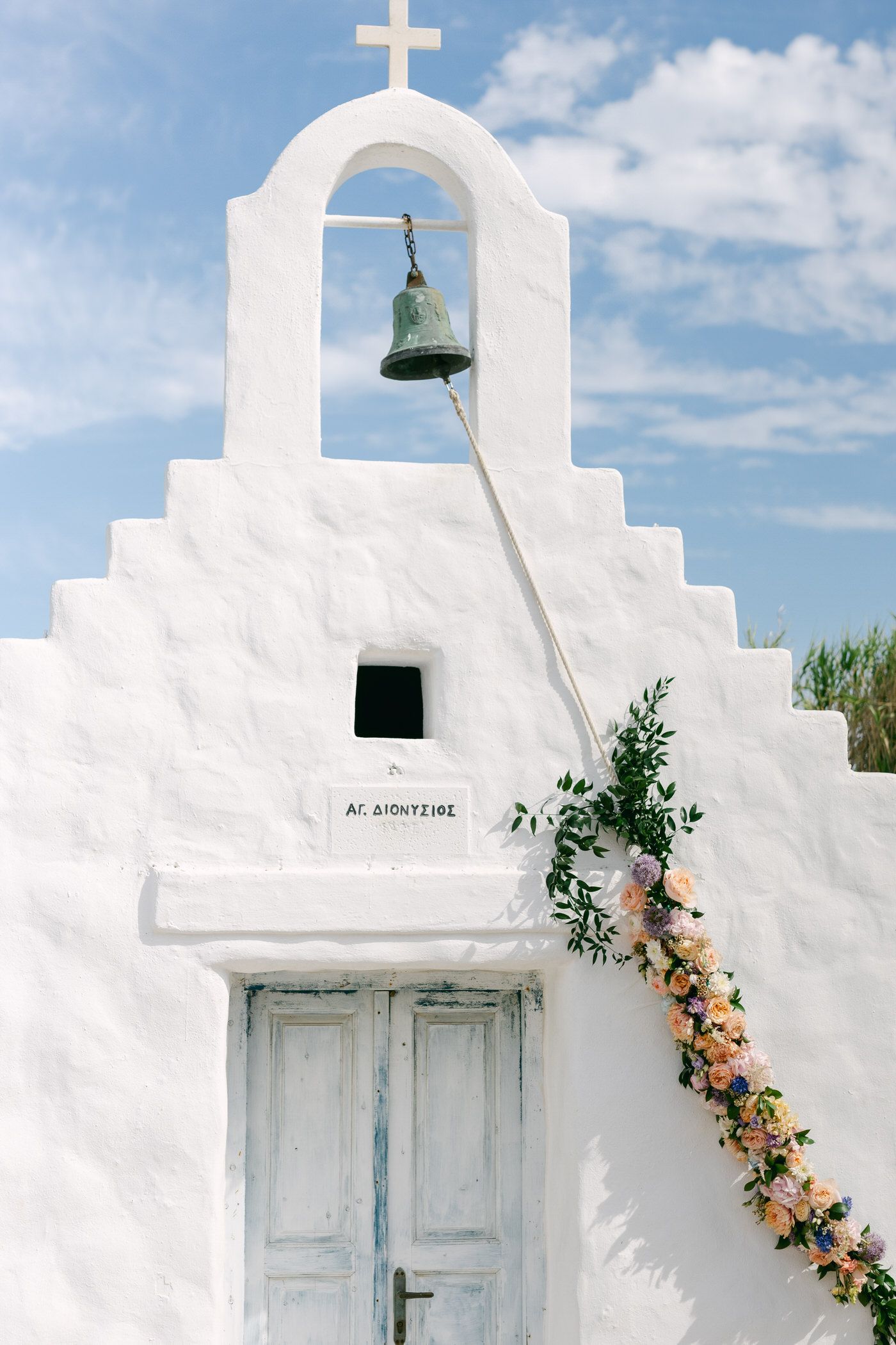 White Mediterranean church facade with bell tower, pale blue door and climbing peach flowers against a blue sky.