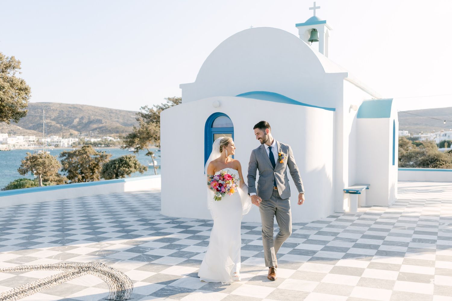 A couple walks hand in hand near a white Mediterranean church with blue dome in Greece.