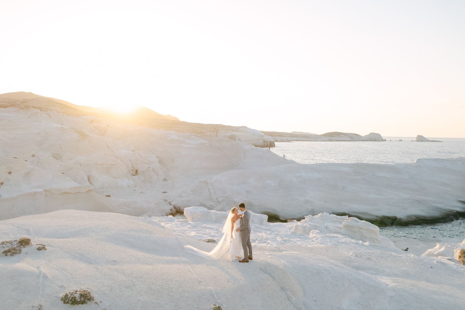 Two figures stand on white coastal rocks at sunset overlooking a dramatic oceanside landscape in Sarakiniko, Greece.