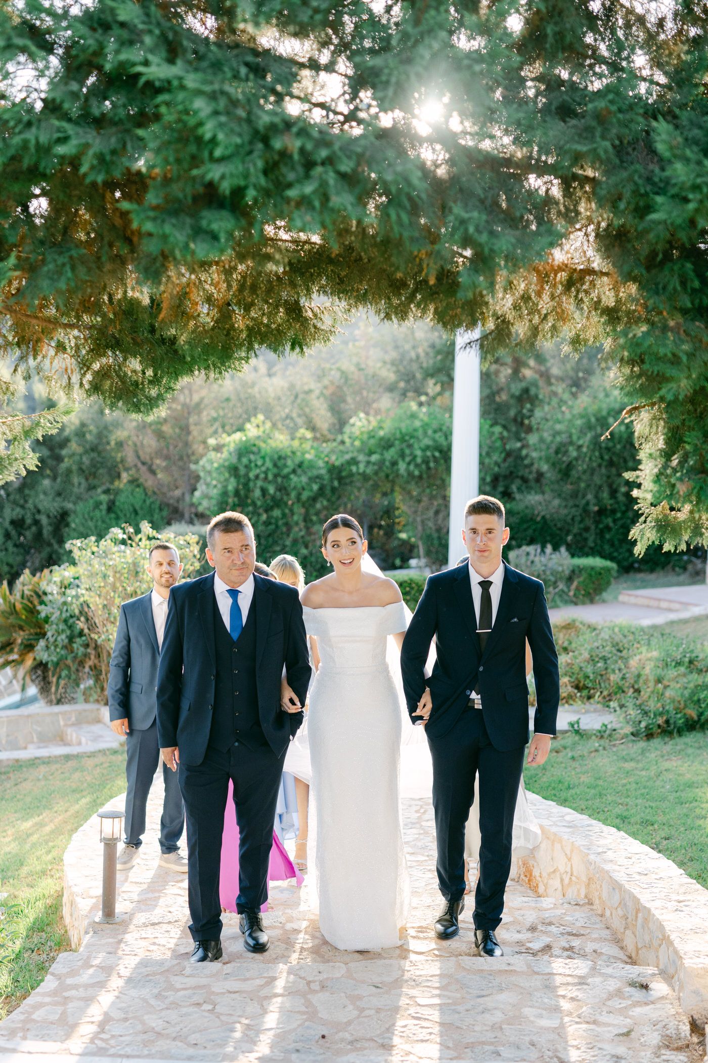 A wedding party walks together along a stone pathway under green trees and golden sunlight in an elegant garden setting.