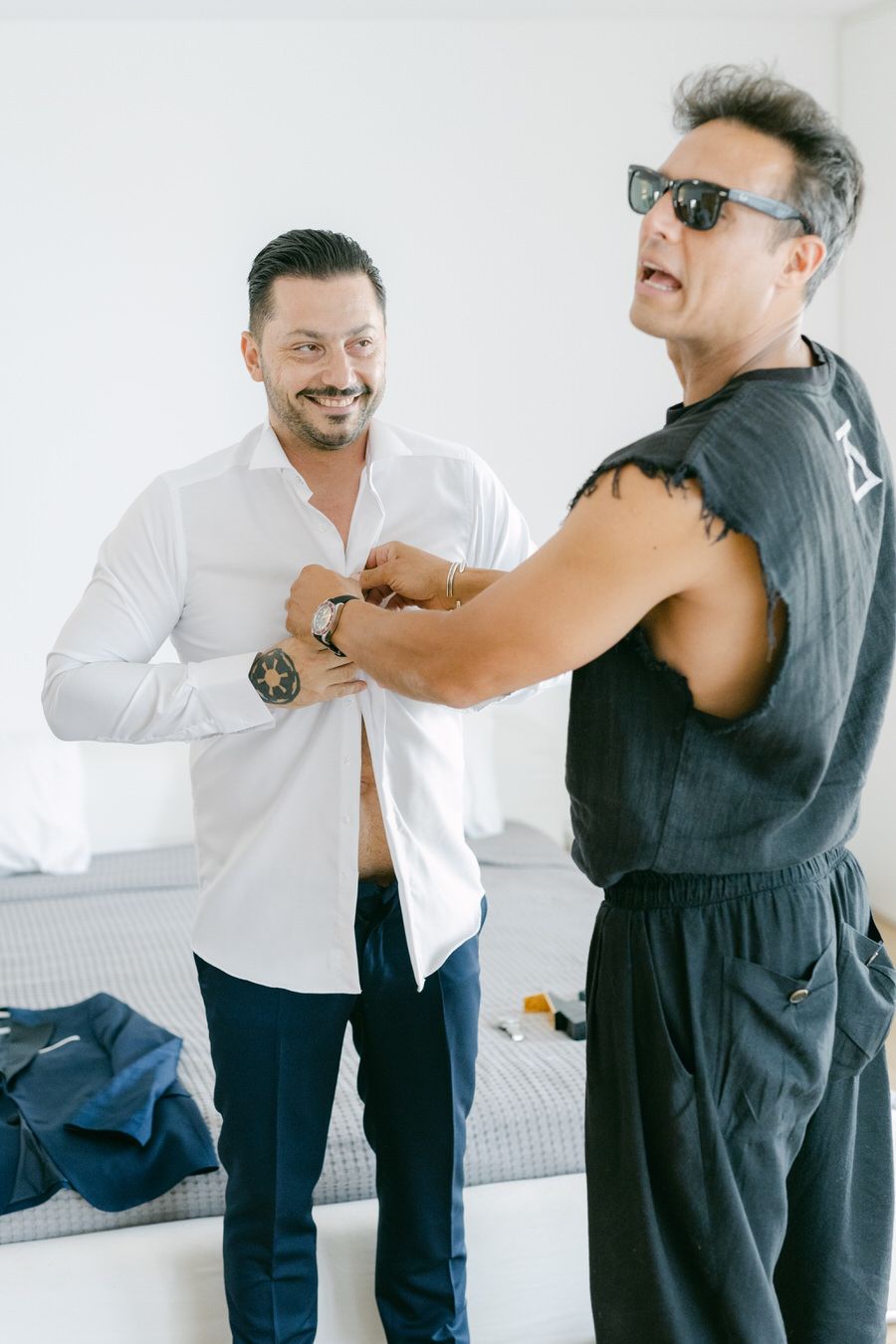 Two men in casual attire getting ready and helping each other with formal accessories.