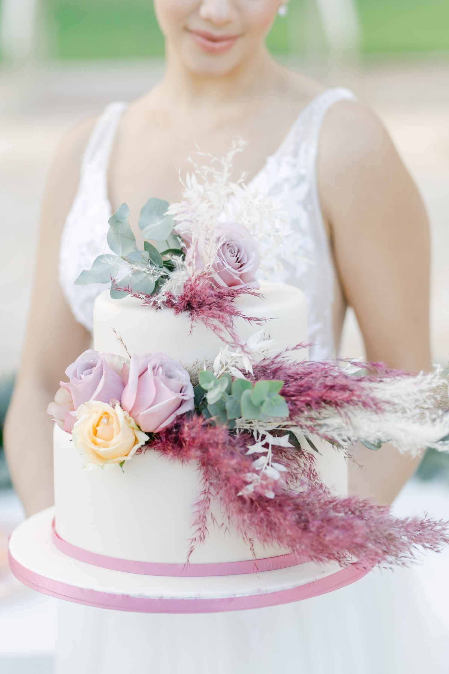 White wedding cake decorated with pink roses, eucalyptus, and burgundy pampas grass.
