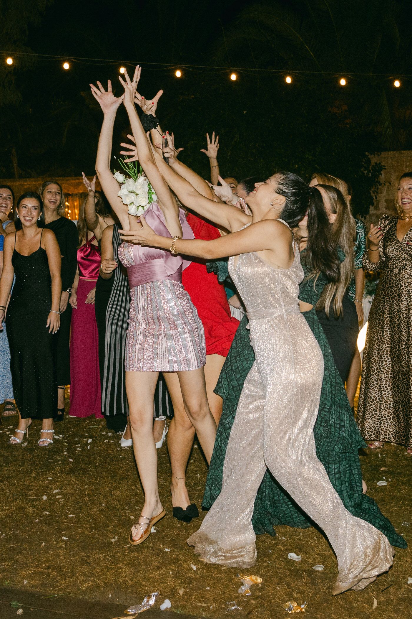 Excited guests in formal dresses jumping and reaching for something at an evening outdoor party with string lights above.