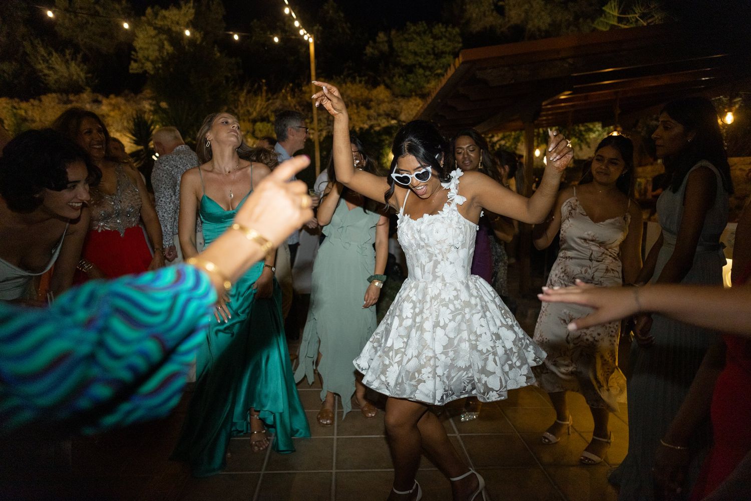 People dancing at a nighttime celebration with string lights overhead and someone in a white lace dress in the center.