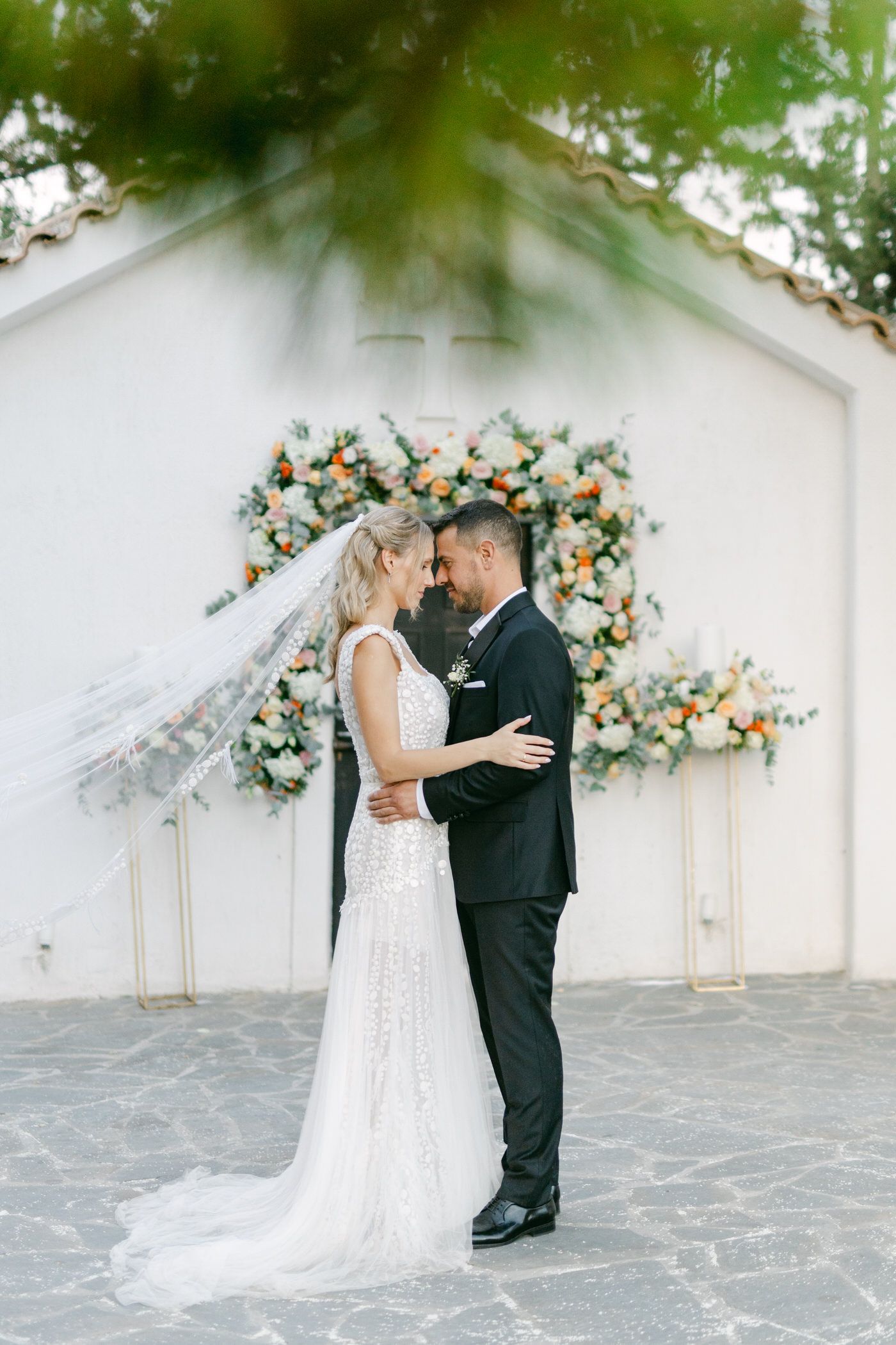 A wedding couple embracing in front of a white wall decorated with peach and turquoise floral arrangements.