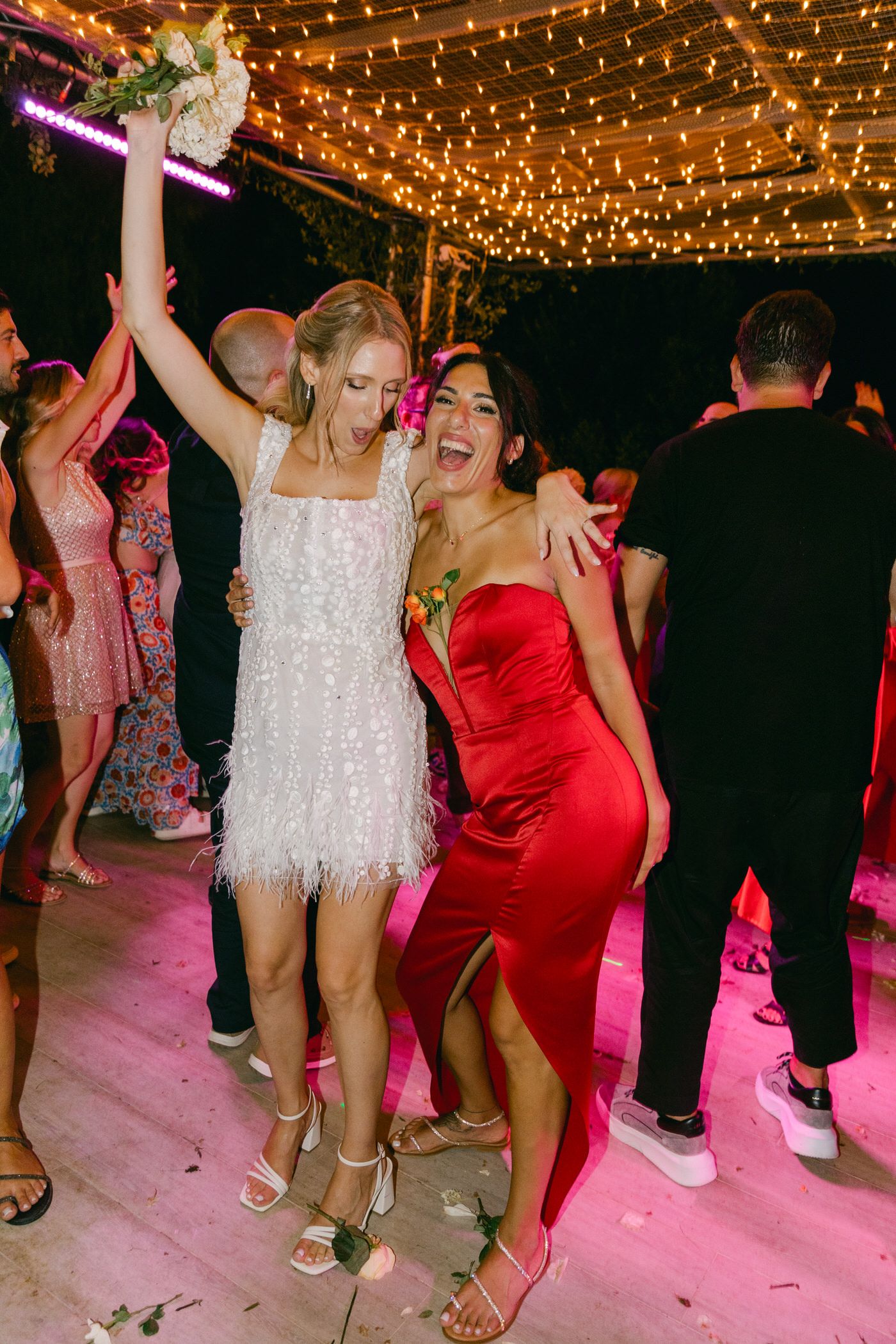 Party guests dance and celebrate under twinkling string lights, one in a sparkly white dress and another in red satin.