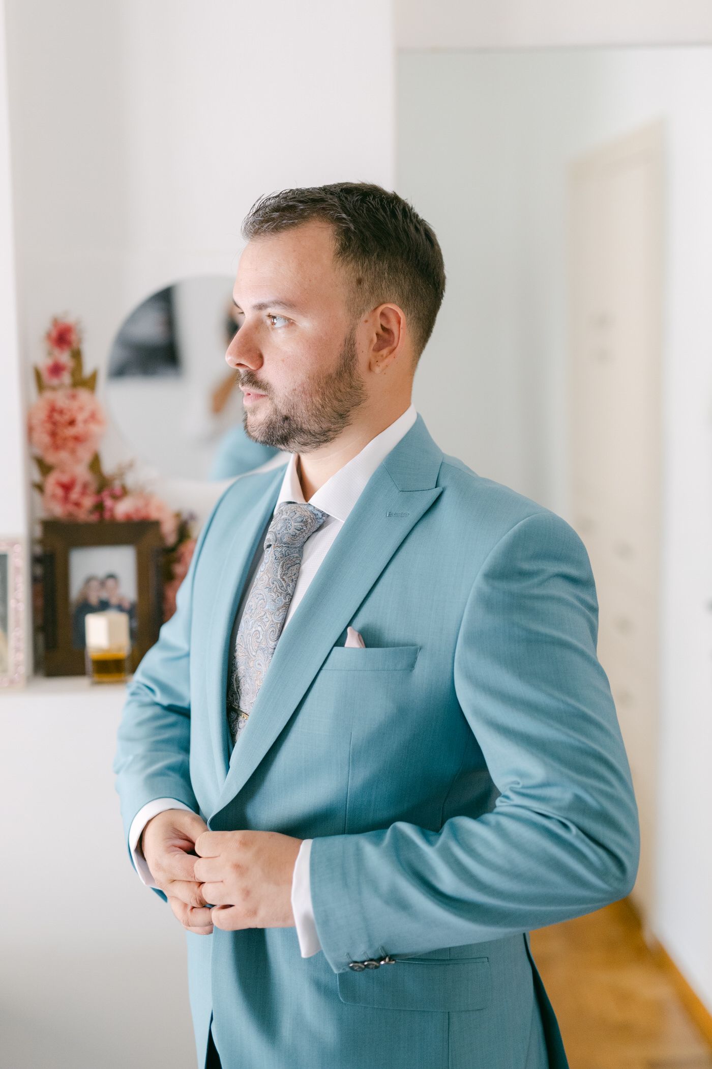 Side profile portrait in a light blue suit and silver tie with elegant decor and flowers in the background.
