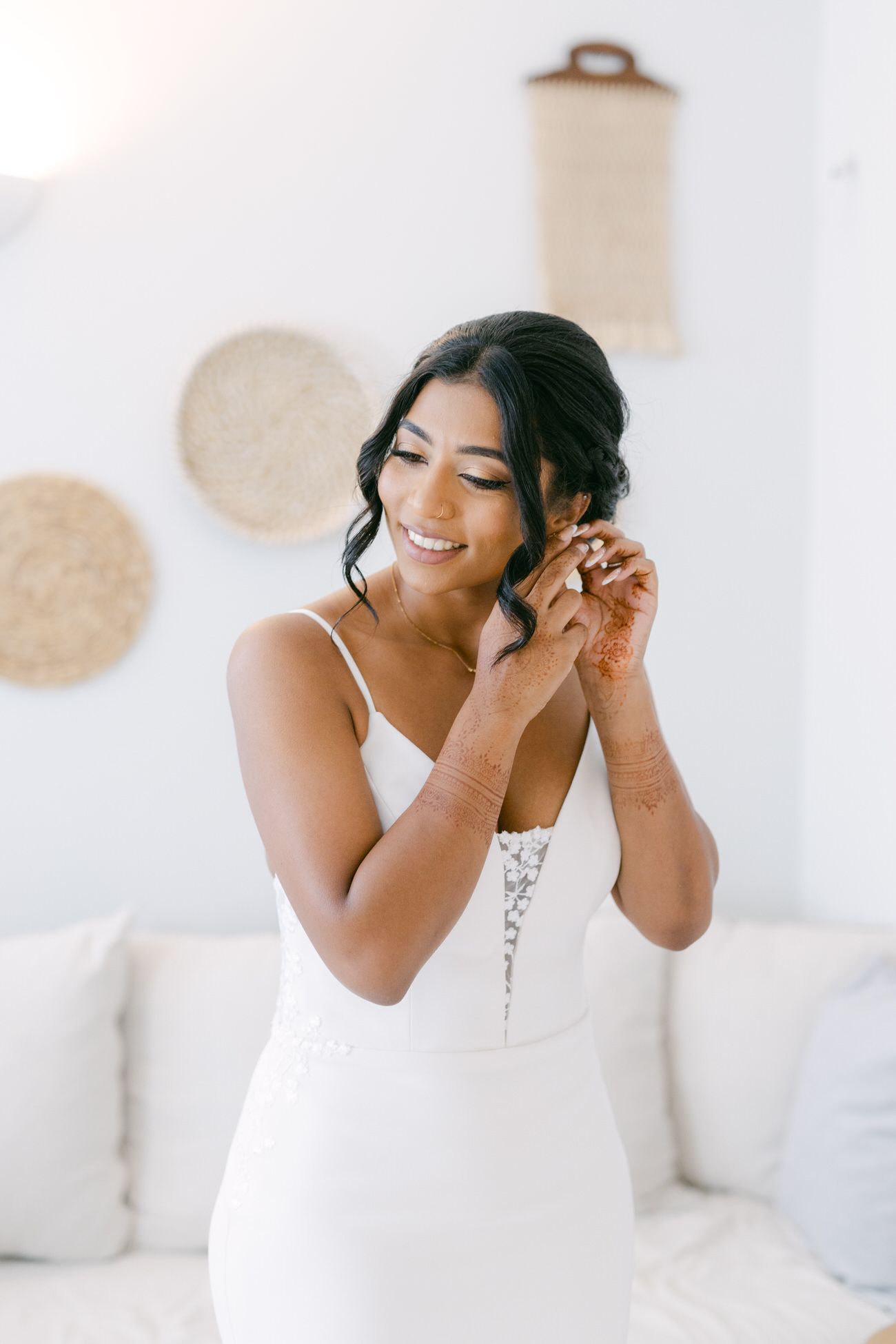 Bride in fitted white dress adjusting earring in bright white room.