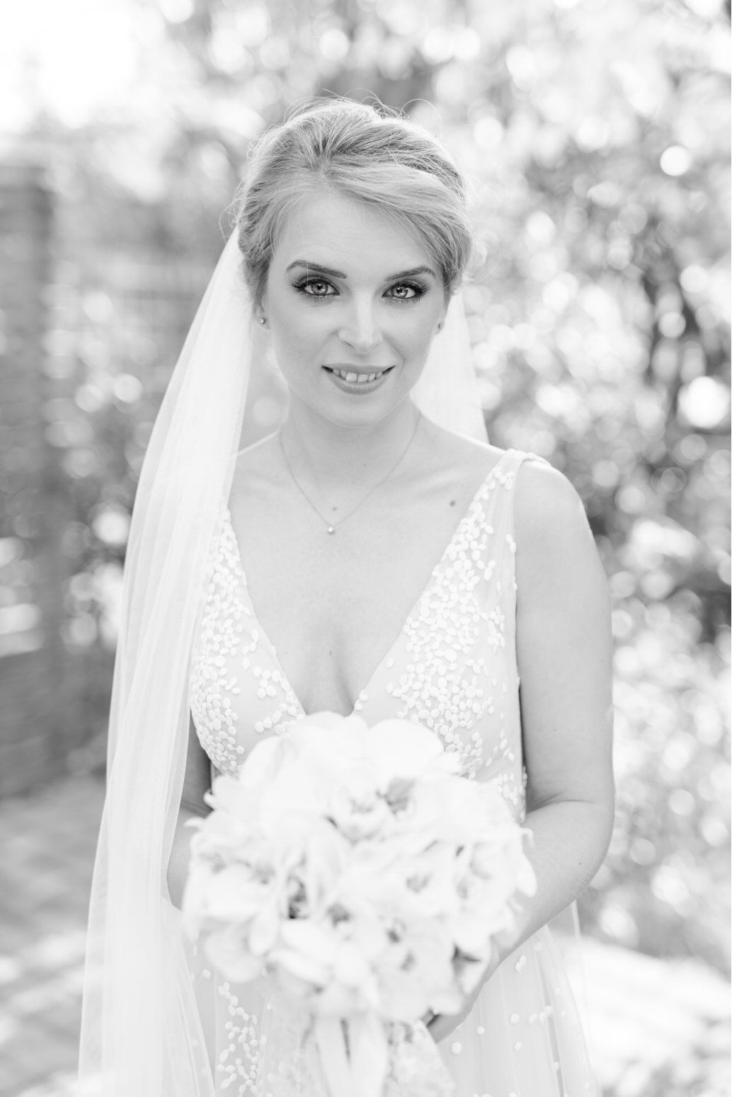 A bride in a white lace wedding dress with a veil holds a white bouquet in this black and white portrait.