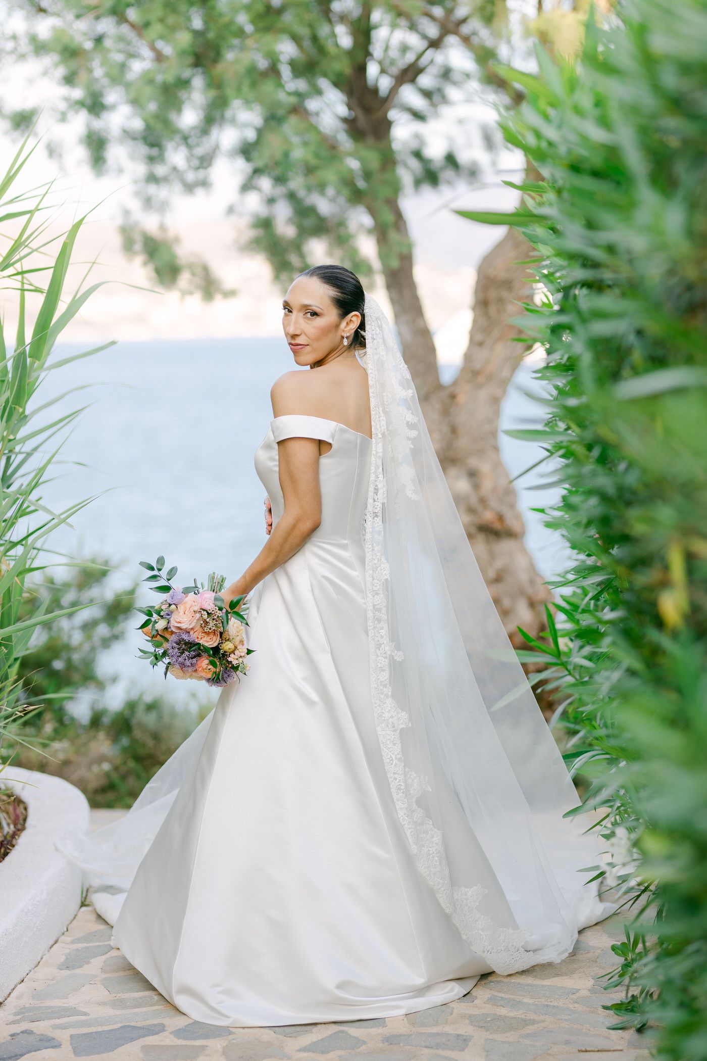 A bride in an elegant off-shoulder white wedding dress and long veil poses with a pink bouquet in a garden setting.