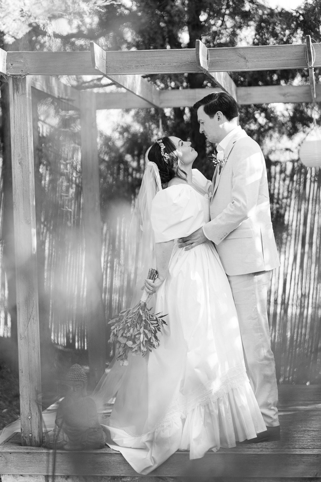 A romantic black and white wedding portrait of a couple embracing under a wooden pergola.