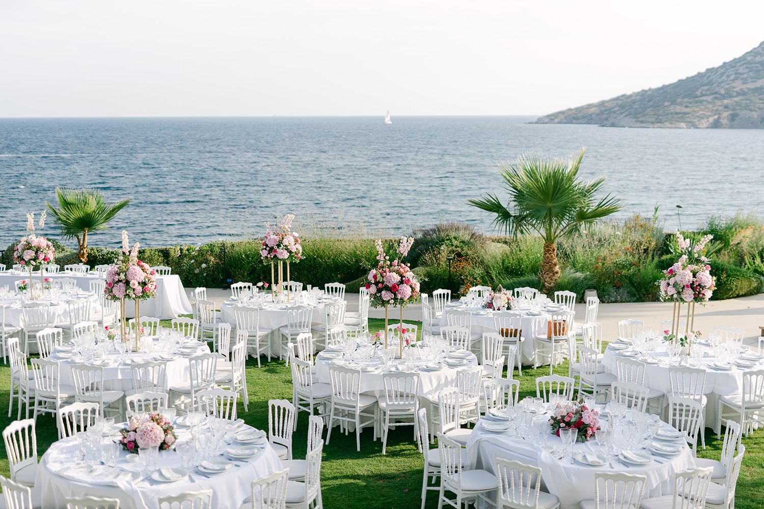 Elegant outdoor wedding reception setup with white tables and chairs overlooking a seaside view with palm trees. Ktima 48. Κτήμα 48.
