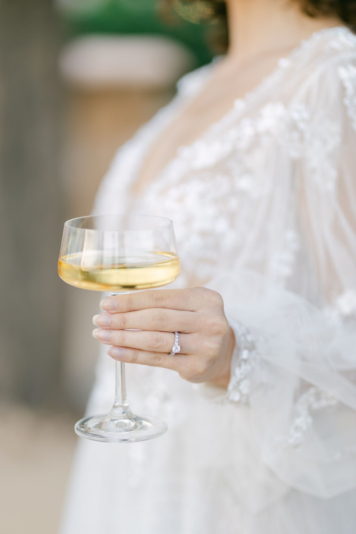 A hand wearing a diamond engagement ring holds a glass of white wine against a white lace dress.